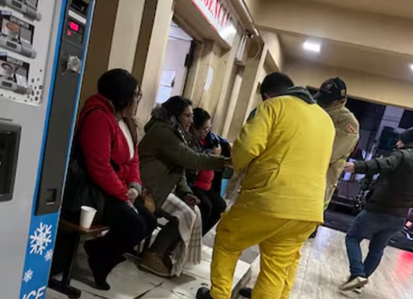 Bomberos voluntarios uniformados distribuyen alimentos calientes a personas sentadas en el interior del Hospital de Ñemby durante la noche, en el marco de su programa solidario de asistencia a vulnerables por las bajas temperaturas.
