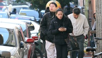 Tres personas caminan por una vereda en una calle céntrica de Asunción, vistiendo camperas gruesas. Una mujer observa su celular mientras un hombre a su lado camina con las manos en los bolsillos. Otro hombre, detrás, lleva una mochila y mira al frente. El entorno urbano muestra vehículos estacionados, bicicletas y un fondo con árboles y más transeúntes, evidenciando una mañana fría y activa.