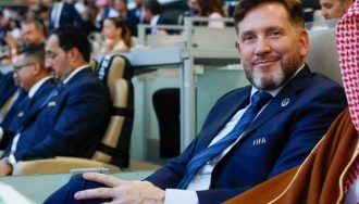 Alejandro Domínguez con traje azul y corbata, con un distintivo de FIFA en la solapa, sentado en una conferencia o evento con una audiencia de fondo. Otros asistentes, también con trajes, están sentados a su alrededor, y una persona con vestimenta tradicional árabe está a su lado.