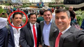 Cuatro hombres vestidos formalmente posan para una selfie en un evento internacional al aire libre, con una multitud y un escenario colorido al fondo. El hombre ubicado a la izquierda de la imagen, señalado con un círculo rojo, destaca por ser el protagonista del contenido relacionado con un nombramiento político. La escena ocurre durante el día, en un ambiente soleado y moderno, con arquitectura contemporánea y presencia de delegaciones internacionales.