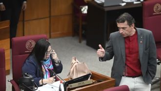 En la imagen se observa a dos senadores paraguayos en el recinto del Senado Nacional. A la izquierda, la senadora Norma Aquino se encuentra sentada en su banca senatorial, sosteniendo un teléfono celular cerca de su oído mientras mantiene una conversación. A la derecha, el senador Juan Carlos Galaverna está de pie junto a su asiento, vestido con un saco gris y camisa roja, haciendo un gesto con su mano derecha levantada mientras parece dirigirse hacia su colega. El ambiente corresponde al hemiciclo del Congreso Nacional, con las características bancas de color bordó y el mobiliario institucional típico del recinto legislativo paraguayo.