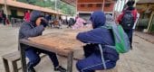 Estudiantes en uniforme azul sentados en bancos de madera bajo una estructura techada en lo que parece ser el patio de una escuela. En el fondo se observan edificios escolares con techos de tejas rojas y otros estudiantes. El ambiente sugiere un recreo o momento de descanso en una institución educativa.