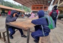 Estudiantes en uniforme azul sentados en bancos de madera bajo una estructura techada en lo que parece ser el patio de una escuela. En el fondo se observan edificios escolares con techos de tejas rojas y otros estudiantes. El ambiente sugiere un recreo o momento de descanso en una institución educativa.