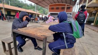 Estudiantes en uniforme azul sentados en bancos de madera bajo una estructura techada en lo que parece ser el patio de una escuela. En el fondo se observan edificios escolares con techos de tejas rojas y otros estudiantes. El ambiente sugiere un recreo o momento de descanso en una institución educativa.