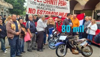 Grupo de jubilados y pensionados municipales manifestándose con pancartas que dicen "PTE. SANTIAGO PEÑA LOS JUBILADOS Y PENSIONADOS MUNICIPALES NO ESTAMOS MEJOR" frente a una motocicleta con el precio "G. 80 mil" superpuesto, en protesta por la venta patrimonial de la Caja Municipal mientras esperan el pago de sus jubilaciones.